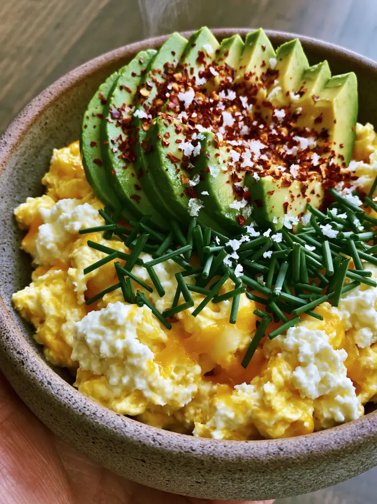 Handheld photo of a vibrant stoneware bowl with soft scrambled eggs and avocado. (breakfast recipes)
