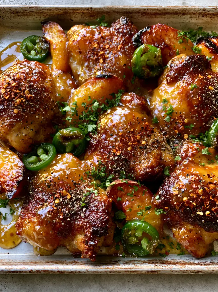Top-down view of glistening chicken thighs with charred peaches and green jalapeño rings on an aluminum tray. (Jalapeño Peach Chicken)