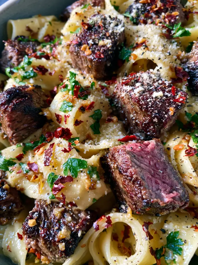 Macro close-up of seared ribeye steak cubes with a juicy pink center over glistening pappardelle pasta. (Cowboy Butter Steak Pasta)