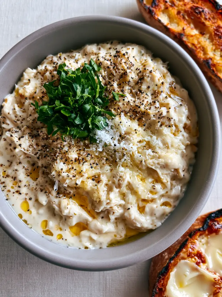 Crockpot Meals: The Ultimate Creamy Garlic Parmesan Shredded Chicken 2 A stone-grey ceramic bowl filled with glossy ivory garlic parmesan chicken and charred sourdough bread representing high-end crockpot meals.