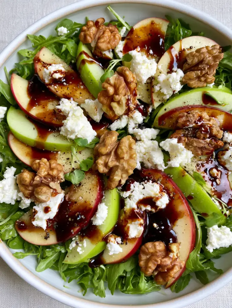 A shallow white ceramic bowl filled with an Apple Arugula Salad, highlighting the glossy maple balsamic vinaigrette and micro-green garnish.
