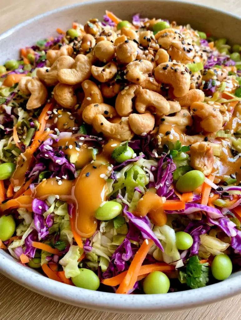 A vibrant crunchy Asian salad in a grey stone bowl featuring purple cabbage, edamame, and a mountain of toasted ramen noodles.