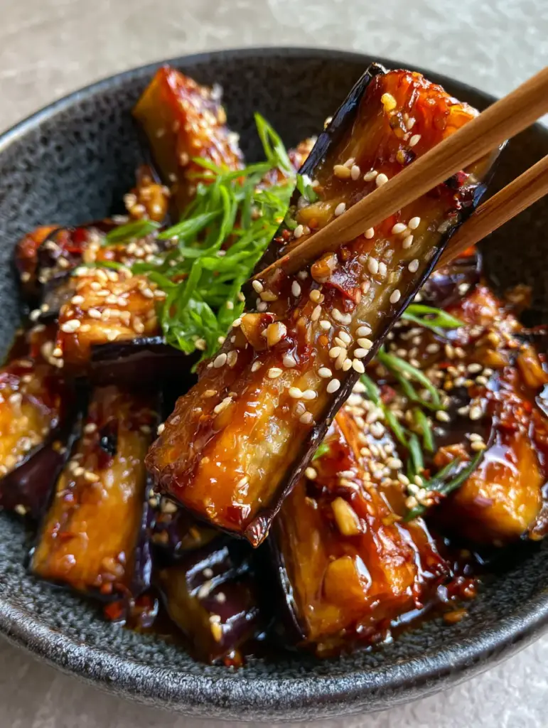 Wooden chopsticks lifting a piece of Sichuan Eggplant showing the creamy white interior and crispy purple skin.