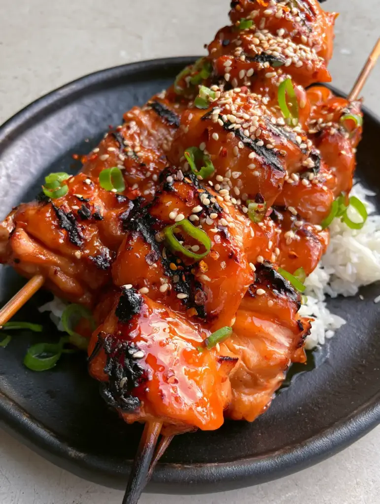 Sweet chili grilled chicken with Sticky Thai Glaze 1 Three skewers of Sweet chili grilled chicken garnished with sesame seeds and green scallions on a black ceramic plate.