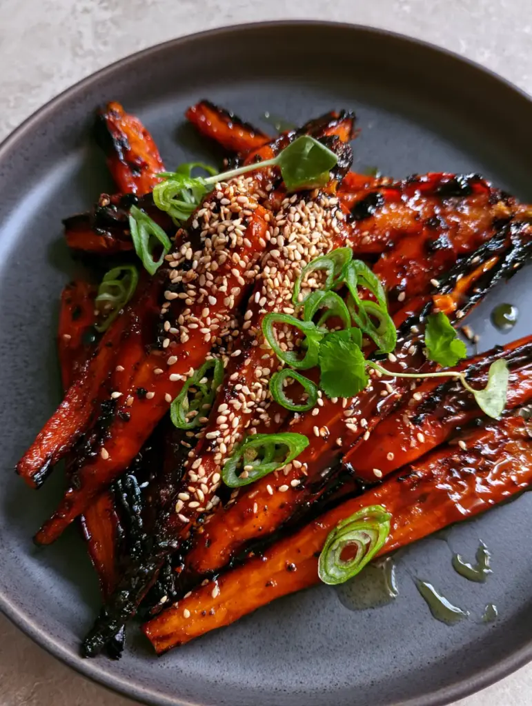 Top-down view of gochujang roasted carrots on a matte plate with cilantro microgreens and lime droplets.
