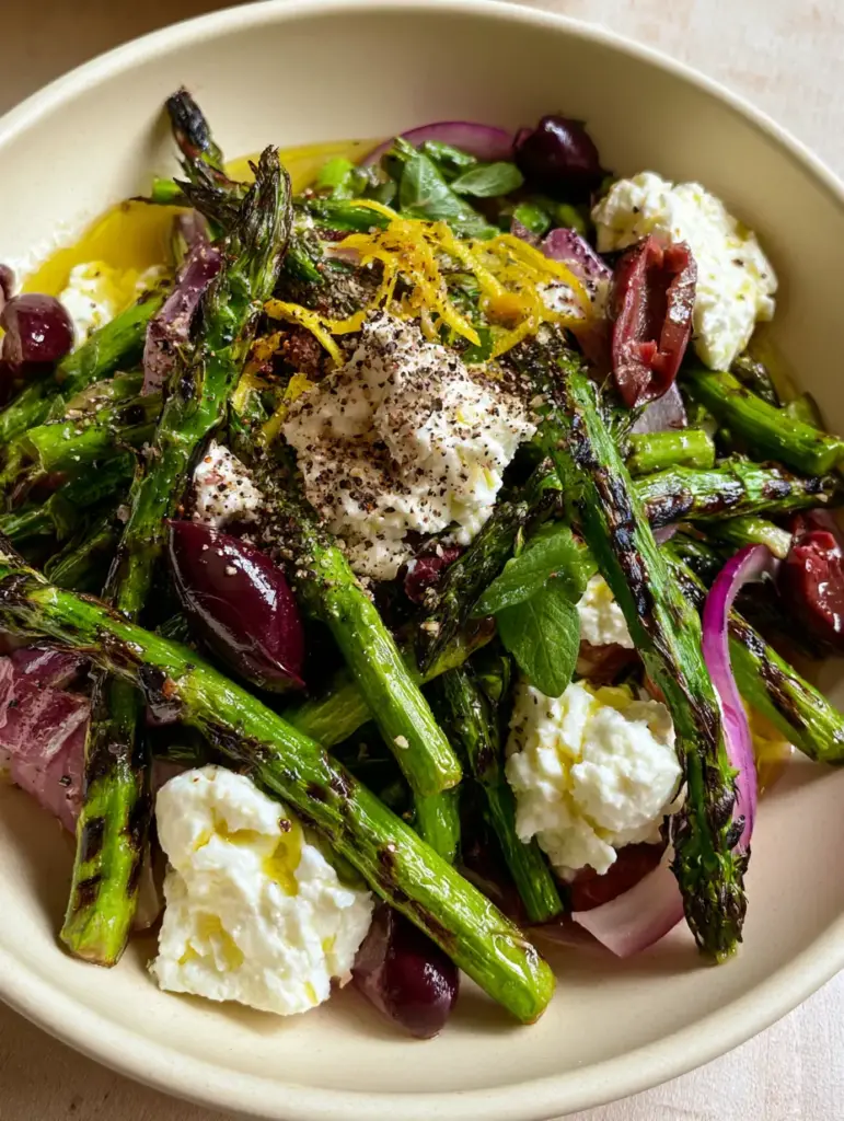 Macro top-down shot of a Greek Asparagus Salad showing the texture of charred asparagus tips, craggy feta, and glossy Kalamata olives.